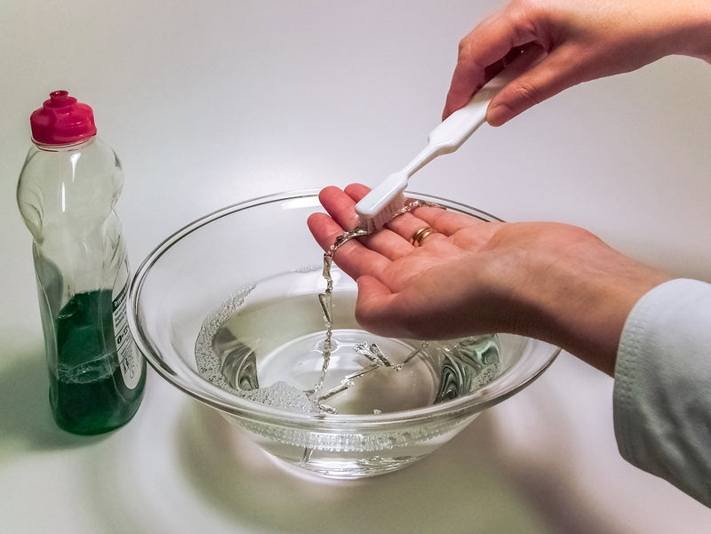 Hands carefully cleaning a silver chain necklace with a soft brush in a glass bowl of soapy water