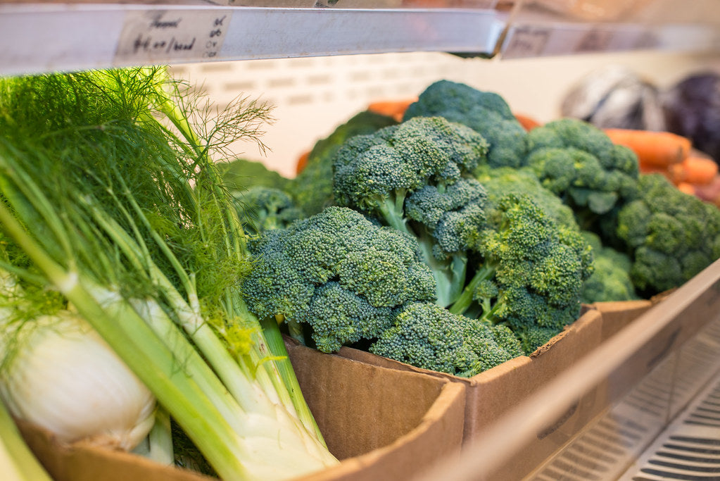 Fresh broccoli, fennel, and carrots in cardboard crates with a handwritten produce price tag at a natural foods market